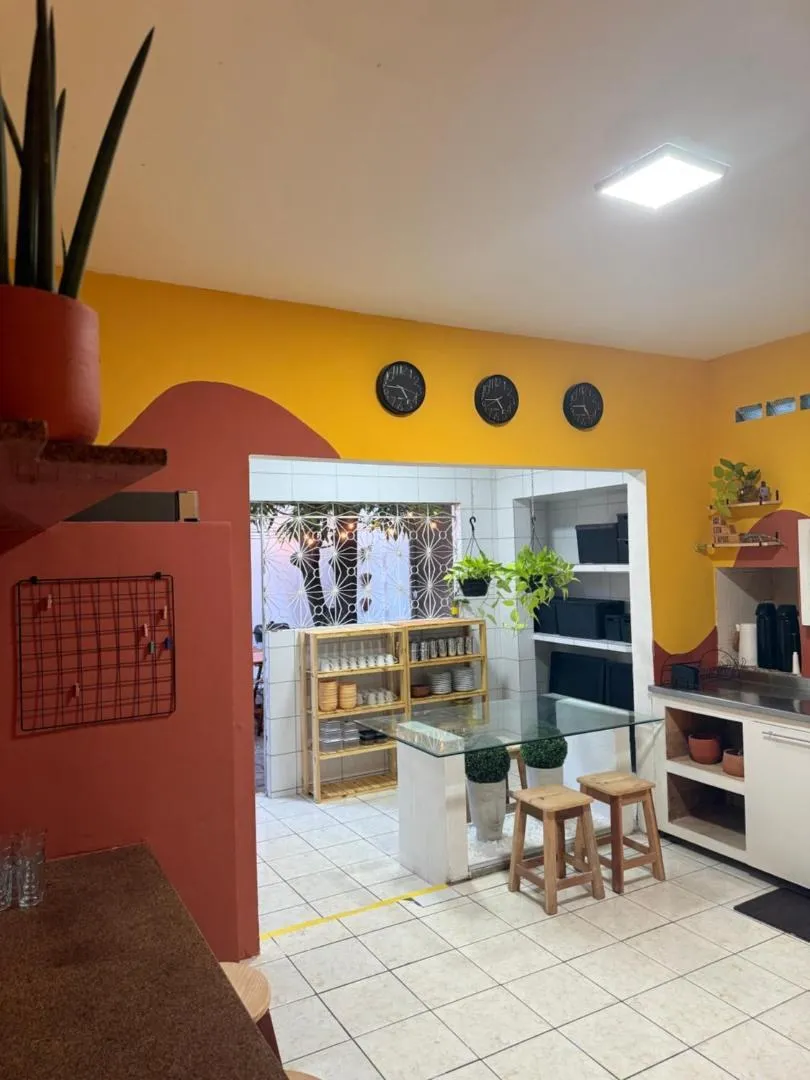 Copa do LumeHostel com paredes amarelas, mural ondulado terracota e mesa de vidro com banquinhos
