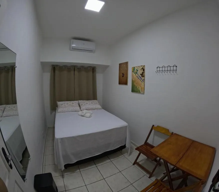 Quarto privativo com cama de casal, cortinas marrons, quadros na parede e mesa com cadeiras